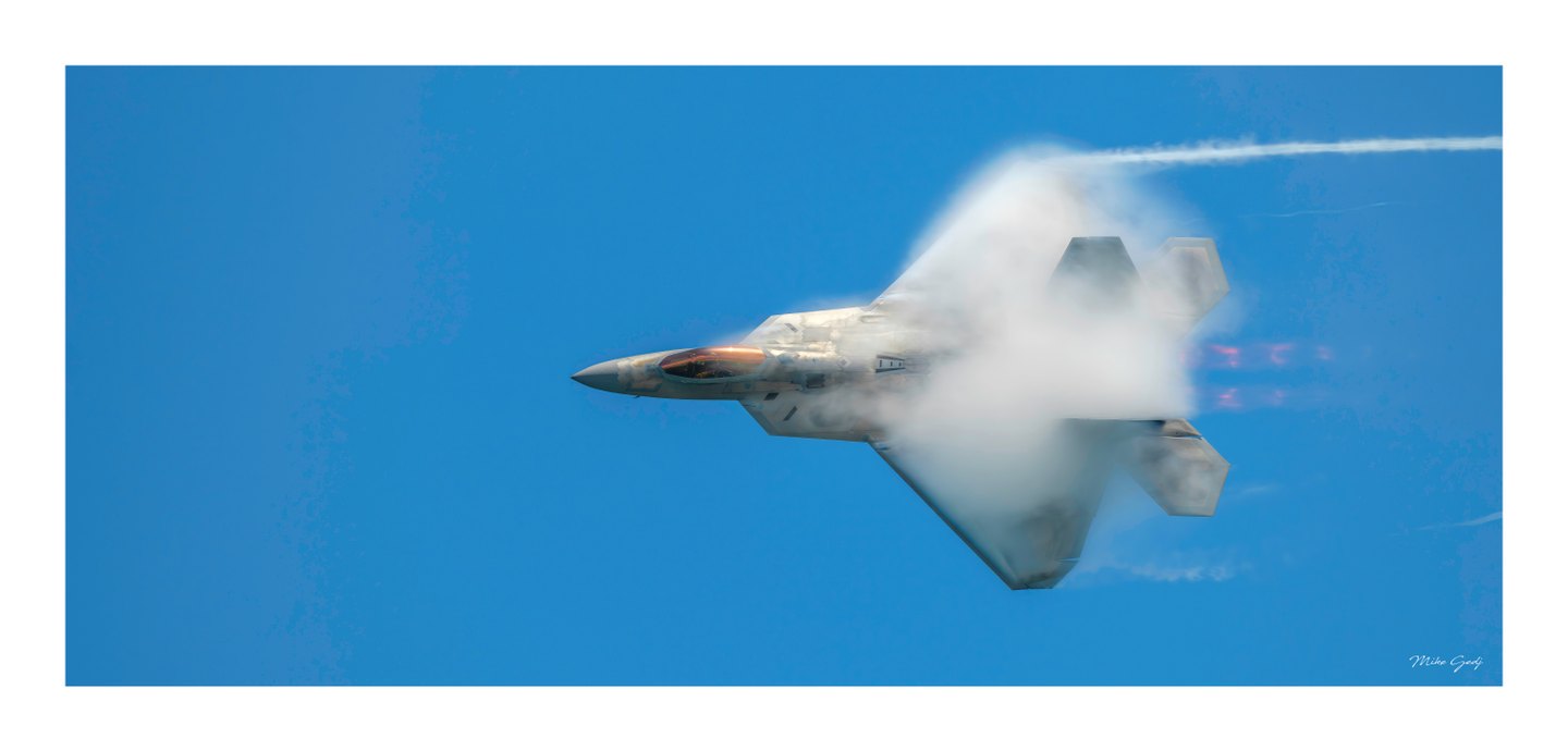 A gray fighter jet banks against a blue sky, its wings partly veiled in white vapor and its canopy gleaming amber.
