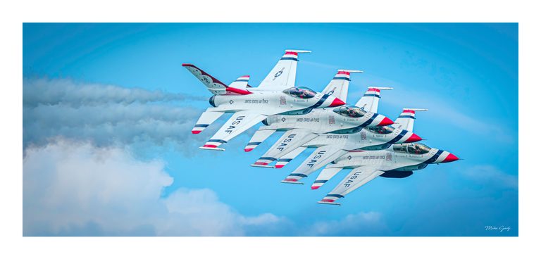 Four white jets with red and blue markings fly in close formation against a blue sky and pale clouds.