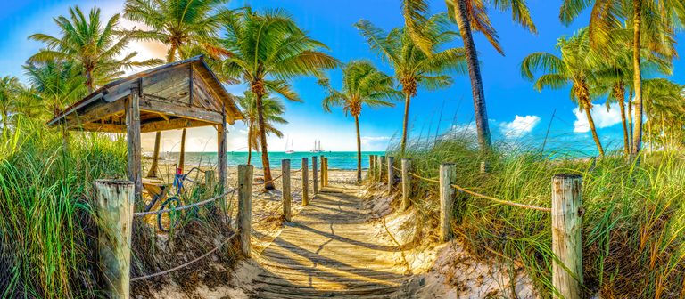 A rope-lined wooden path passes dune grasses and a weathered shelter toward palms, turquoise sea, and two sailboats.
