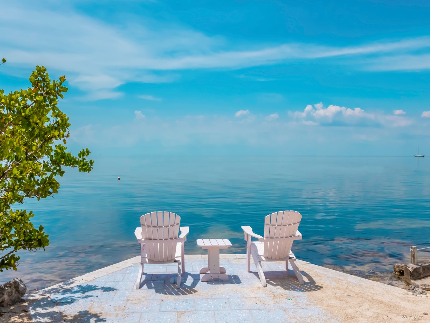 Two white chairs and a small table face calm turquoise water, with leafy branches at left and a distant sailboat.