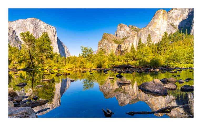 Sunlit cliffs and green forest reflect in calm, rock-strewn water beneath a clear blue sky.