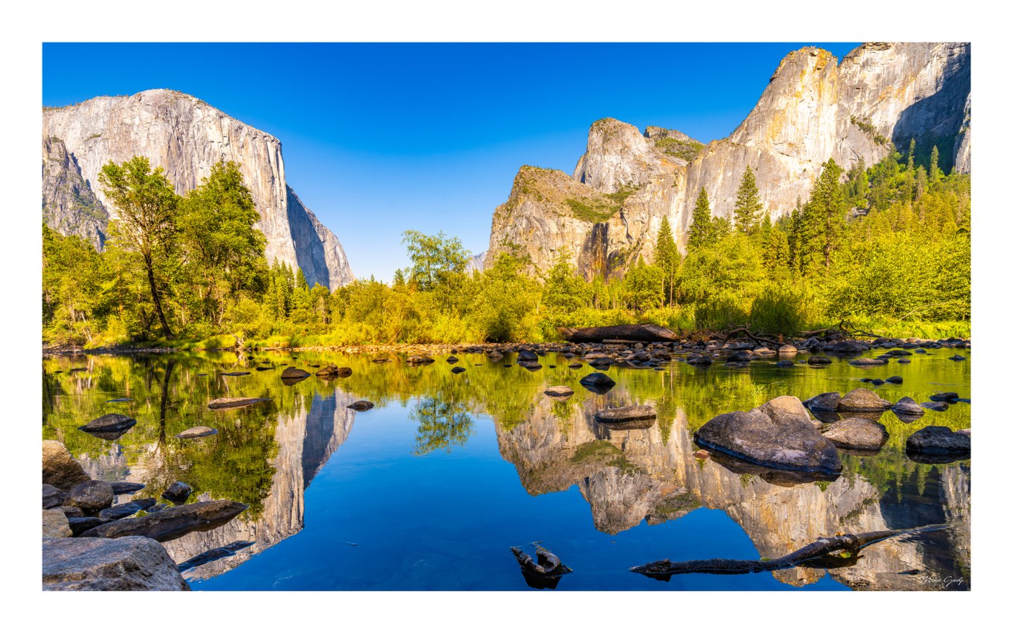 Sunlit cliffs and green forest reflect in calm, rock-strewn water beneath a clear blue sky.