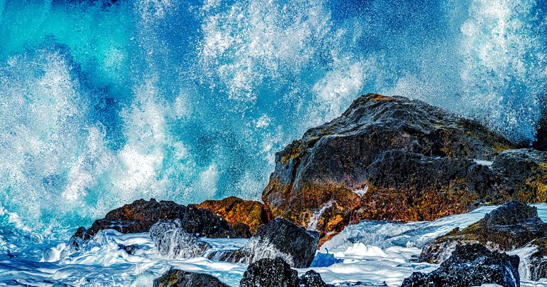 Turquoise surf bursts into white spray above dark, jagged rocks, with foam pooling across the foreground.