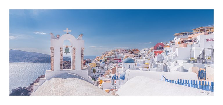 A white bell arch overlooks sunlit sea, rugged cliffs, and hillside buildings with blue domes and pastel walls.