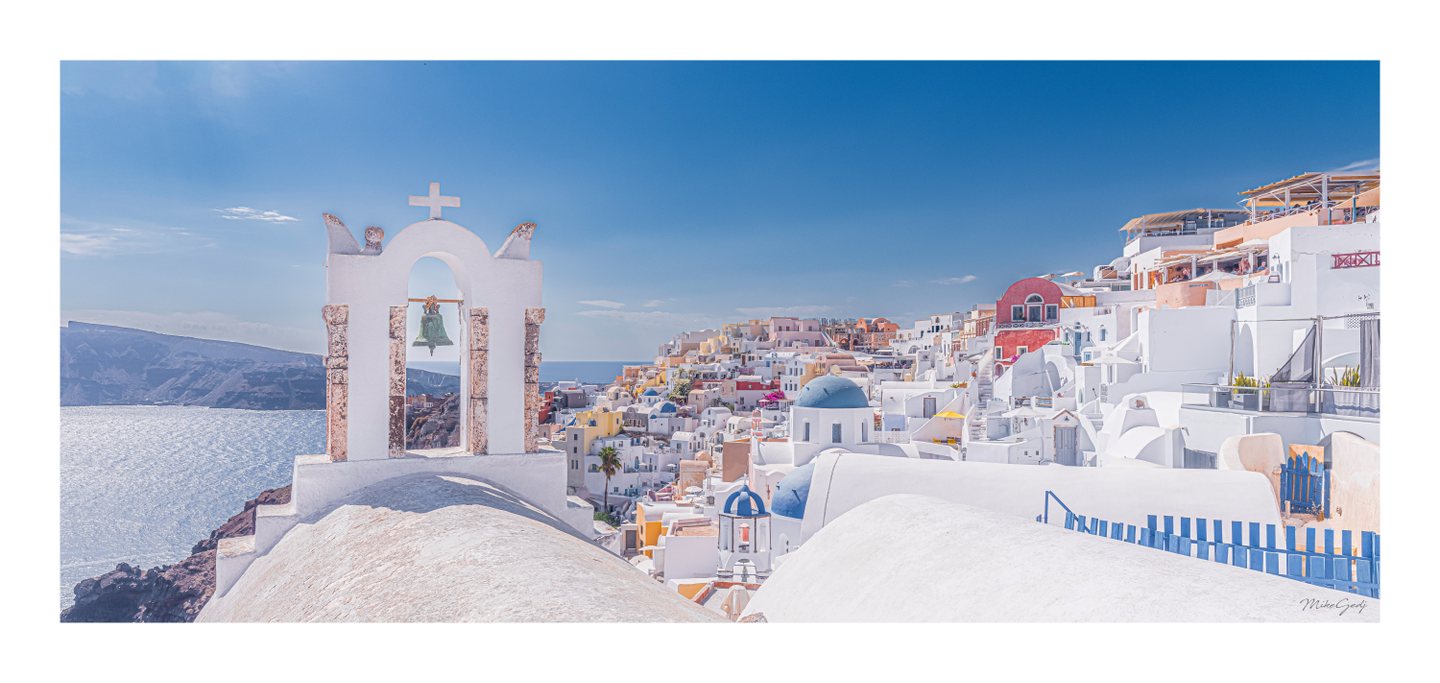 A white bell arch overlooks sunlit sea, rugged cliffs, and hillside buildings with blue domes and pastel walls.