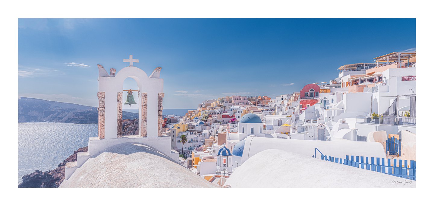 A white bell arch overlooks sunlit sea, rugged cliffs, and hillside buildings with blue domes and pastel walls.