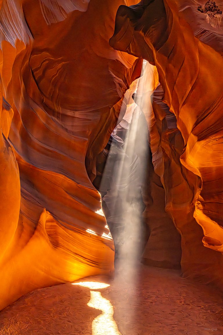 A shaft of sunlight falls between curved orange sandstone walls onto a sandy canyon floor marked with footprints.