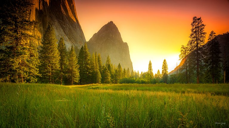 A green meadow and tall conifers stand beneath sheer cliffs and a pointed rock peak under an orange sky.