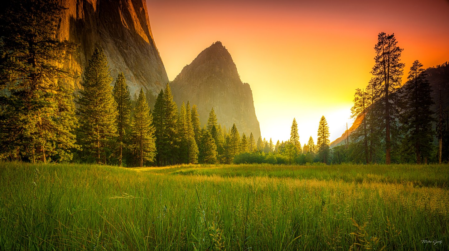 A green meadow and tall conifers stand beneath sheer cliffs and a pointed rock peak under an orange sky.