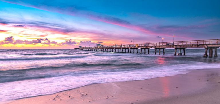A lit pier extends over rolling surf beneath pink and blue clouds, with wet sand reflecting the colorful sky.