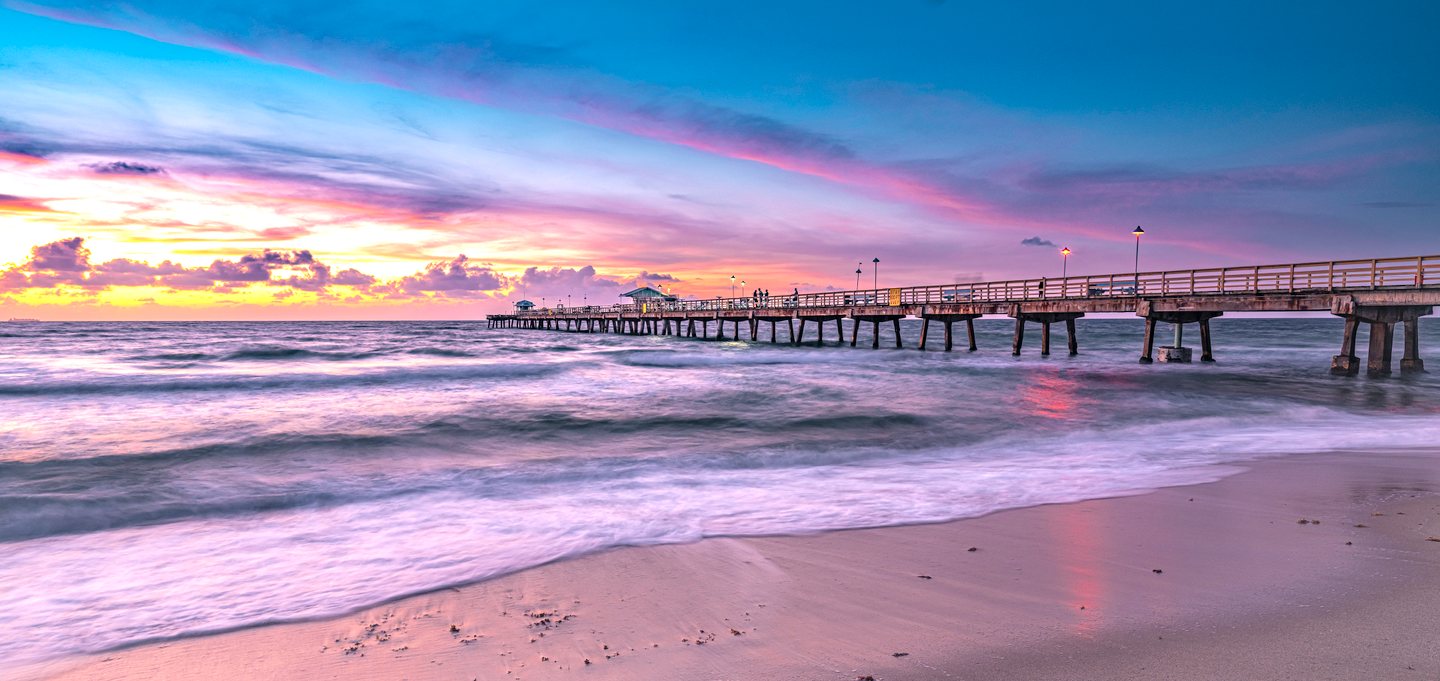 A lit pier extends over rolling surf beneath pink and blue clouds, with wet sand reflecting the colorful sky.