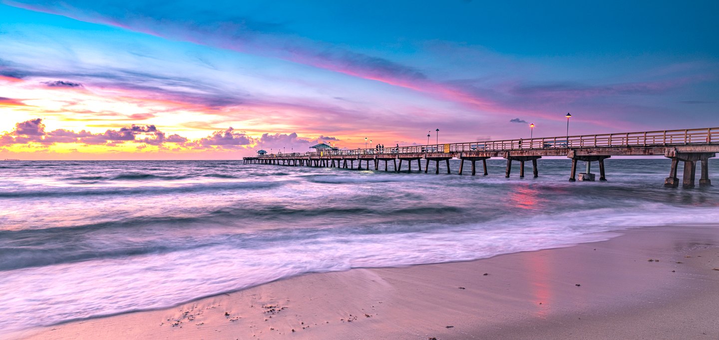 A lit pier extends over rolling surf beneath pink and blue clouds, with wet sand reflecting the colorful sky.