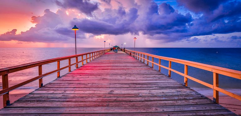 A wooden pier with glowing lamps stretches across calm water beneath pink, purple, and blue clouds.