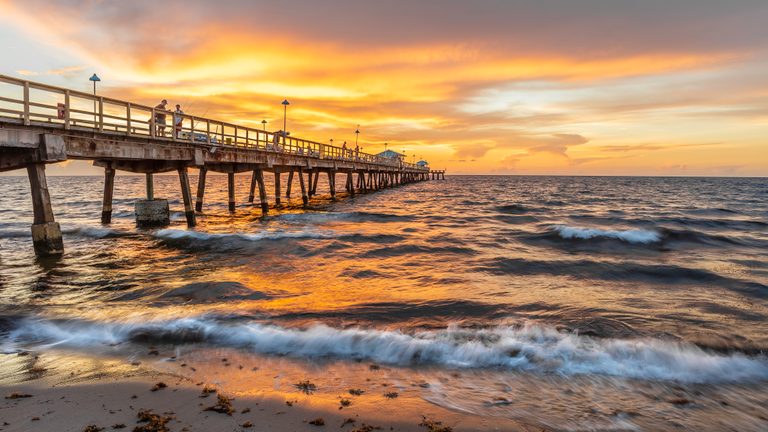 Anglers stand on a long pier above rolling waves beneath an orange sky, with golden reflections on wet sand.