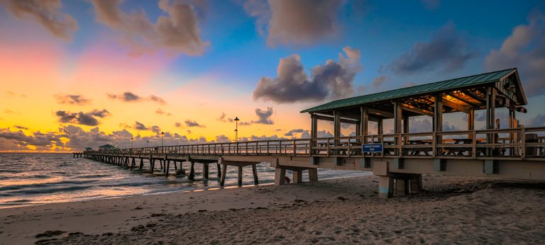 A fishing pier with a lit pavilion stretches over waves beneath gold and pink clouds beside a footprint-covered beach.