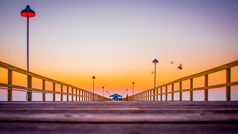 Low view along a wooden pier with yellow railings, glowing lamps, distant people, and birds against an orange sky.