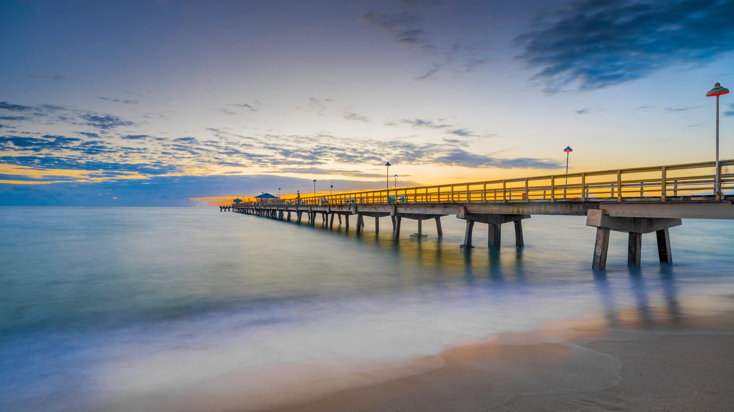 A lamp-lined pier stretches over smooth blue water beneath golden clouds, with wet sand in the foreground.