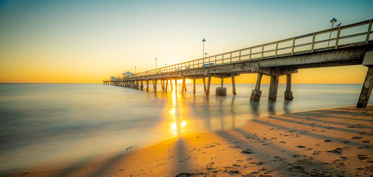 Low sunlight shines beneath a long concrete pier, reflecting across smooth seawater and footprint-marked sand.