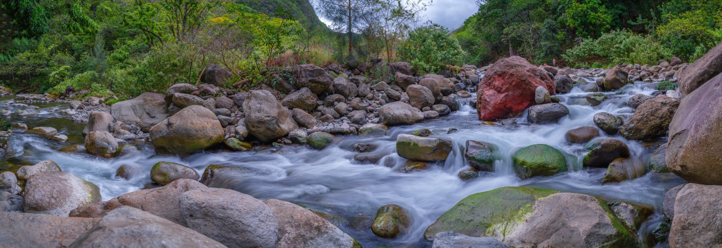 A forest stream cascades among large gray and red boulders, with green moss and small figures on the far bank.