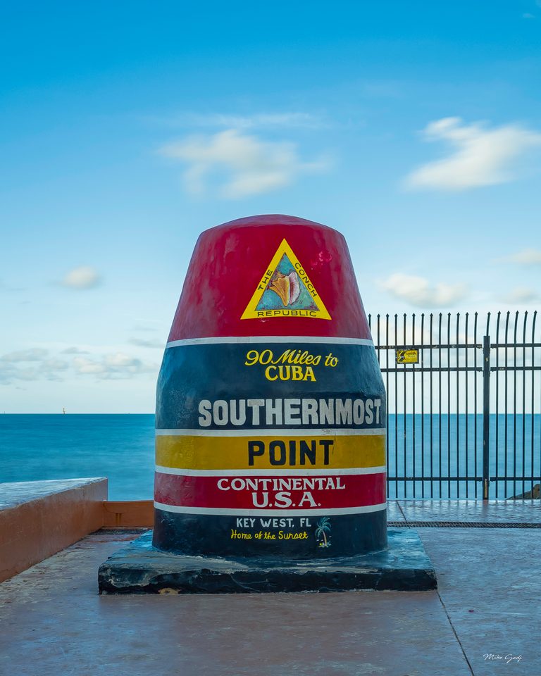 Key West’s striped Southernmost Point marker stands beside a black fence, with turquoise sea and blue sky behind it.