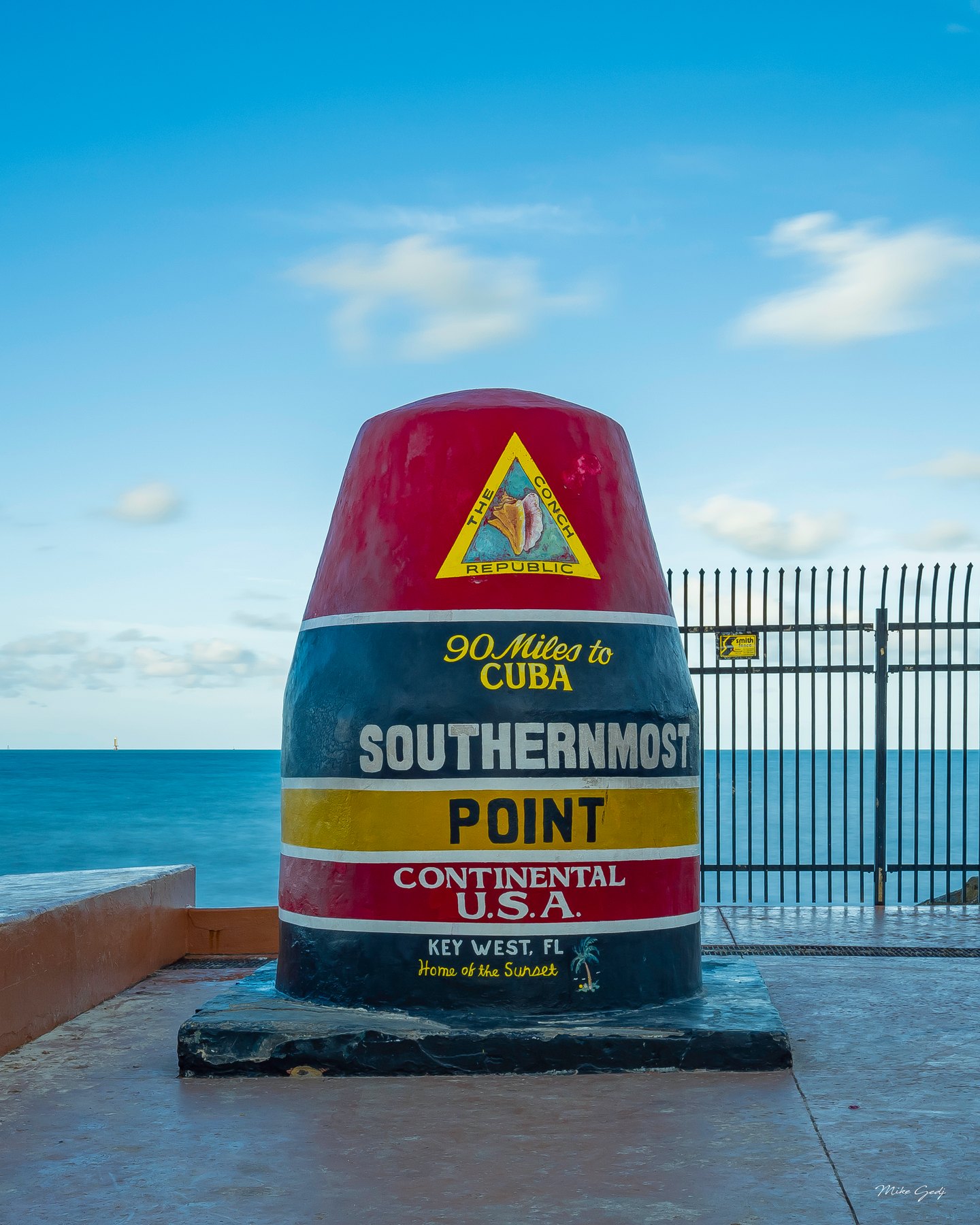 Key West’s striped Southernmost Point marker stands beside a black fence, with turquoise sea and blue sky behind it.