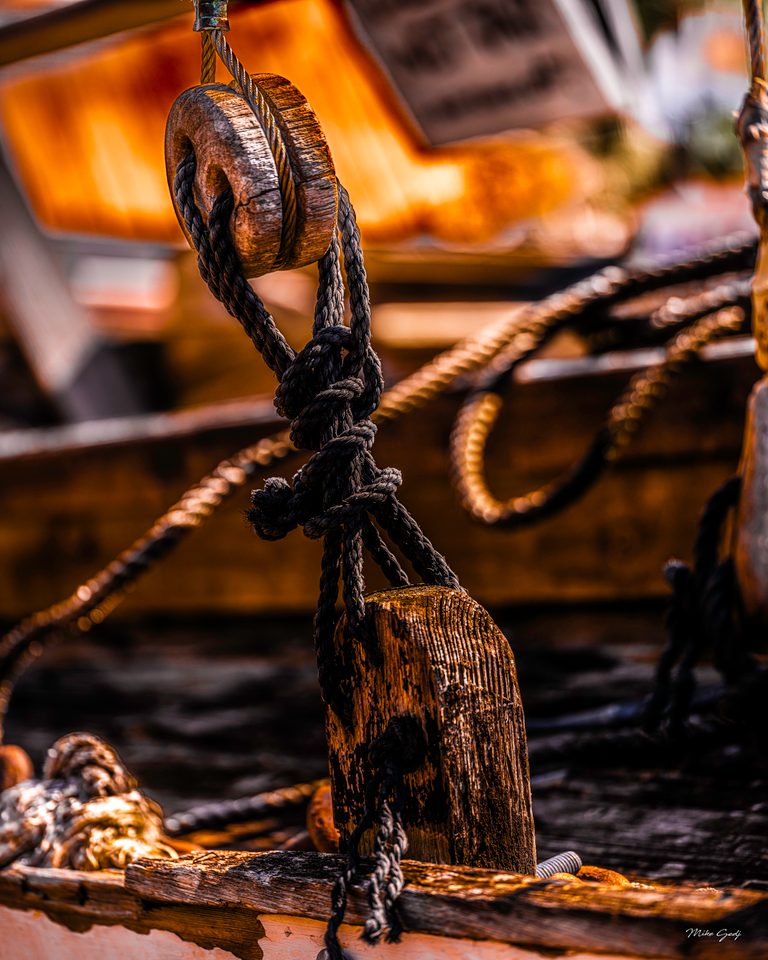 Dark rigging ropes thread through weathered wooden fittings against a softly blurred golden background.