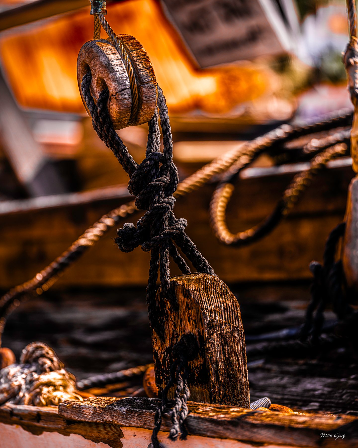 Dark rigging ropes thread through weathered wooden fittings against a softly blurred golden background.