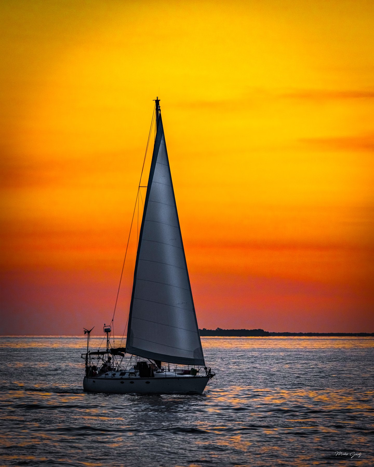 A sailboat with a tall raised sail crosses rippled water beneath an orange sky and a distant dark shoreline.