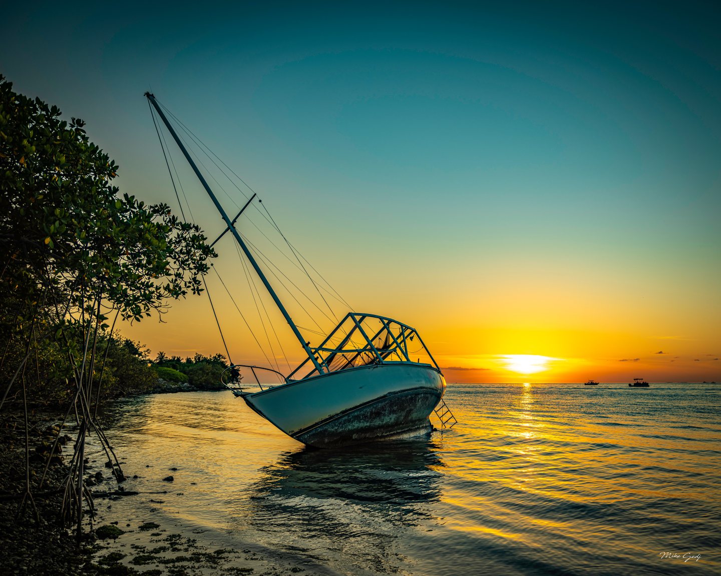 A weathered sailboat leans in shallow water beside mangroves, with a golden sunset and small boats offshore.