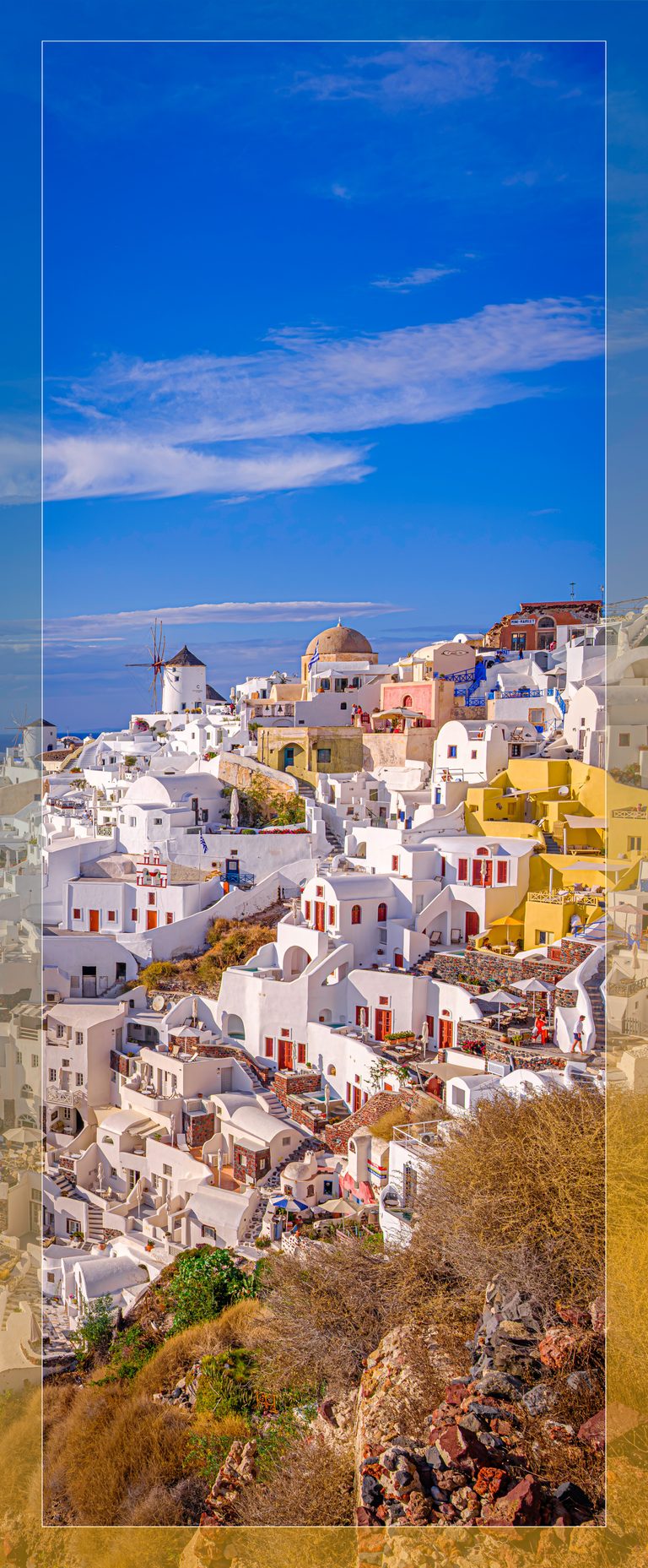 White hillside buildings, windmills and winding stairs rise above dry brush beneath a blue sky streaked with clouds.