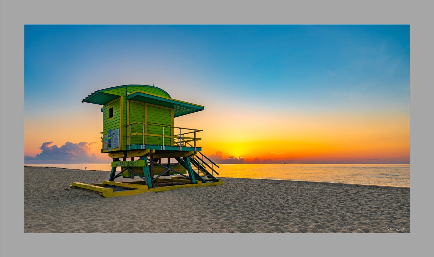 A green and yellow Miami Beach lifeguard tower stands on sand beside a calm ocean beneath a blue and orange sky.