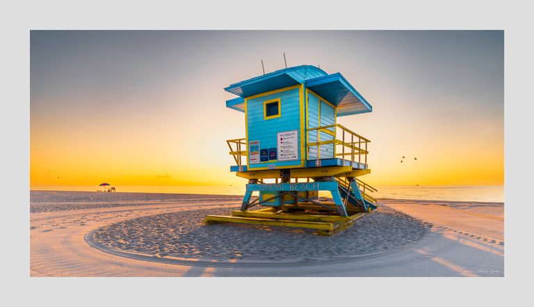 A turquoise and yellow Miami Beach lifeguard tower stands on rippled sand beside a calm sea under a golden sky.