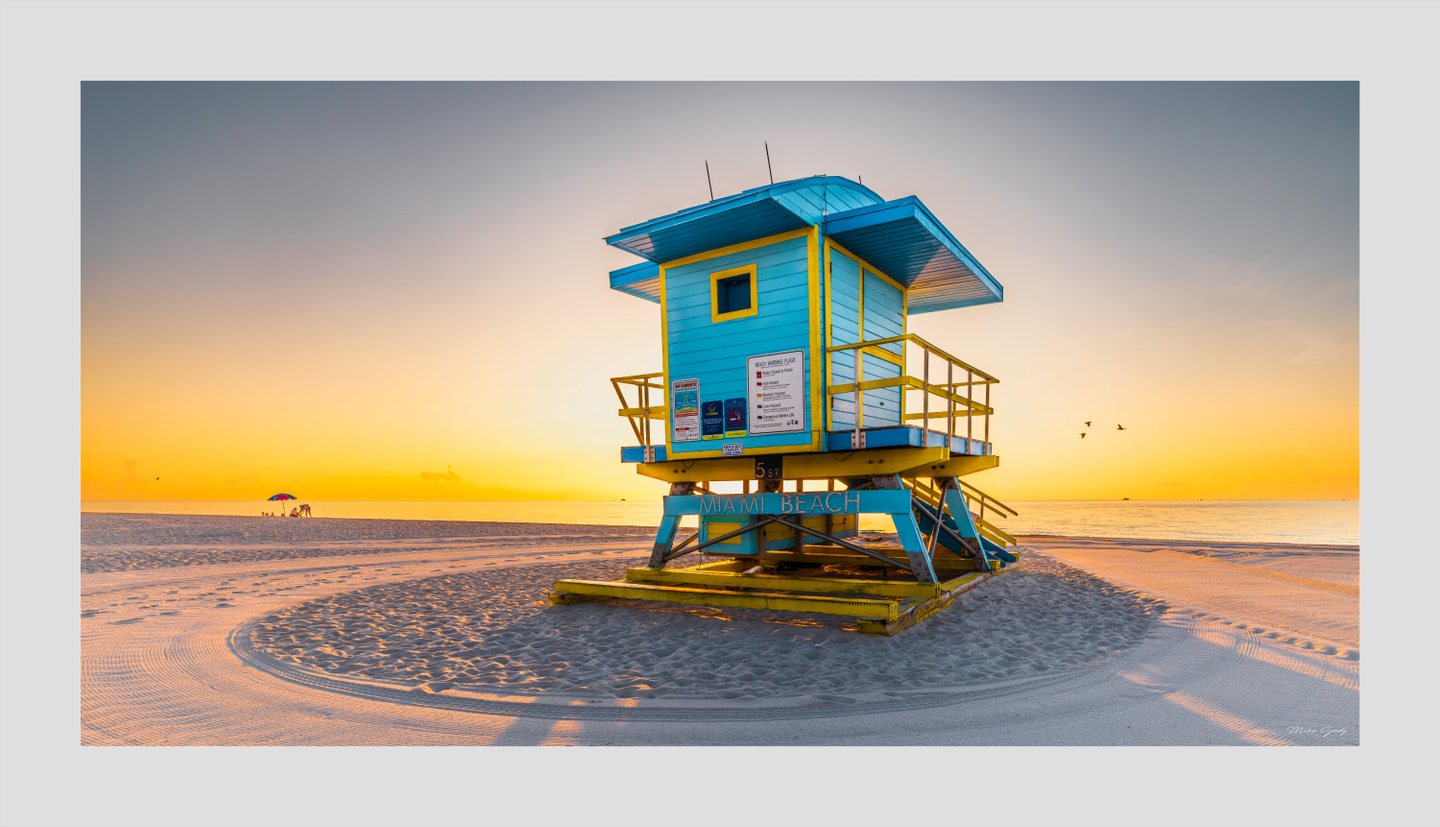 A turquoise and yellow Miami Beach lifeguard tower stands on rippled sand beside a calm sea under a golden sky.