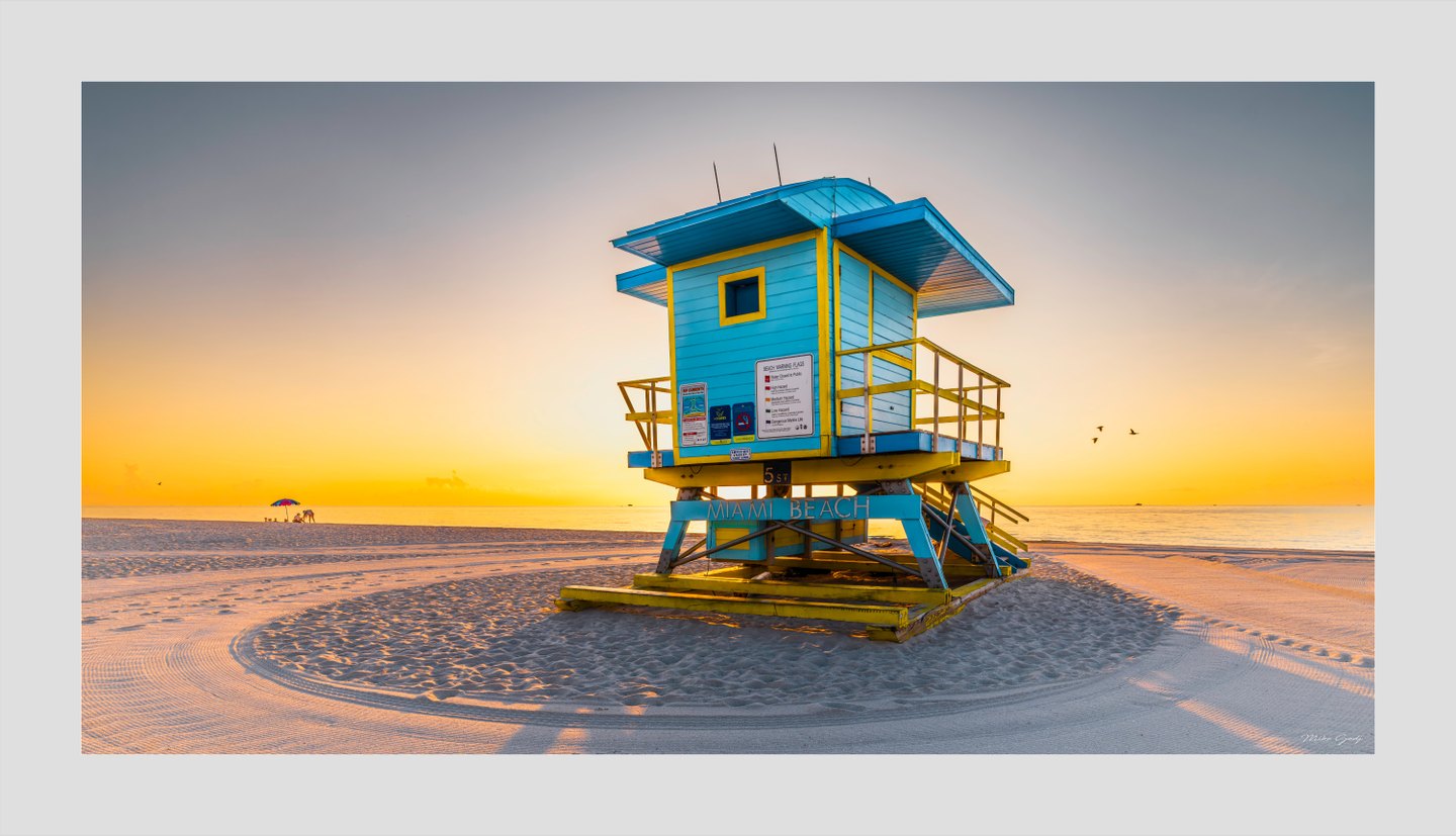 A turquoise and yellow Miami Beach lifeguard tower stands on rippled sand beside a calm sea under a golden sky.