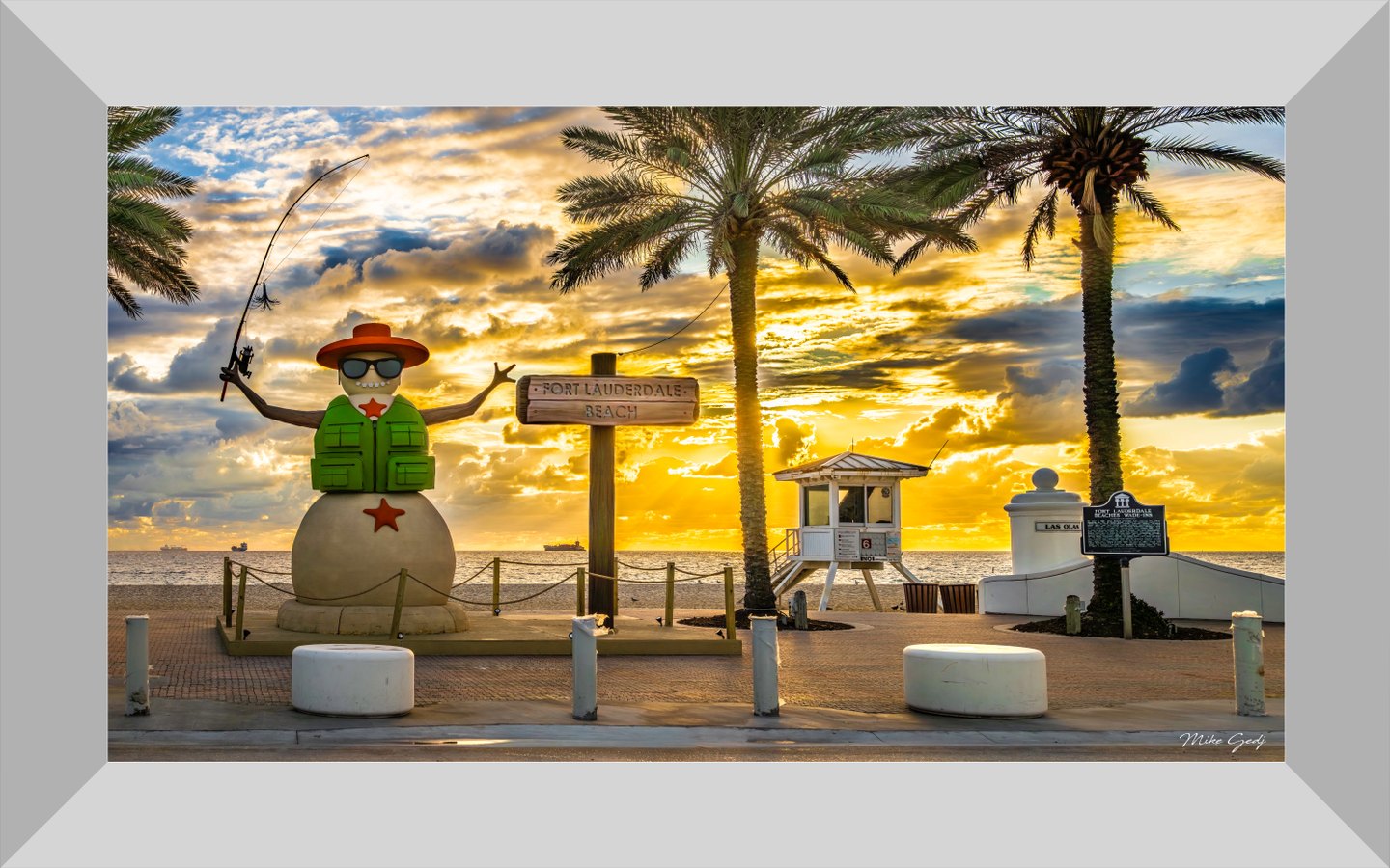 A snowman fishing figure stands beside a Fort Lauderdale Beach sign, palms and a lifeguard tower beneath golden clouds.