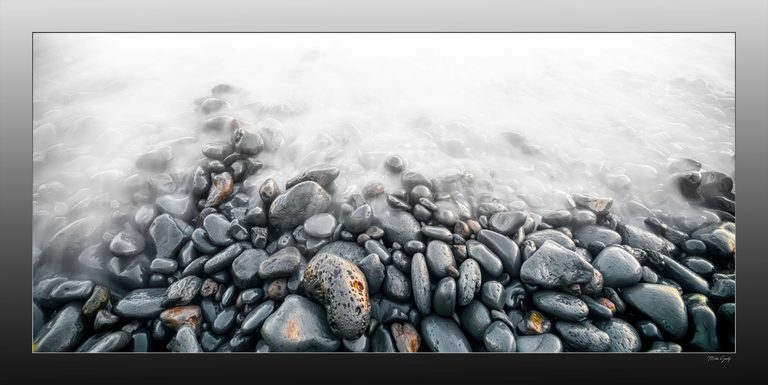 Wet gray pebbles with scattered rust-colored markings emerge from pale, softly blurred water.