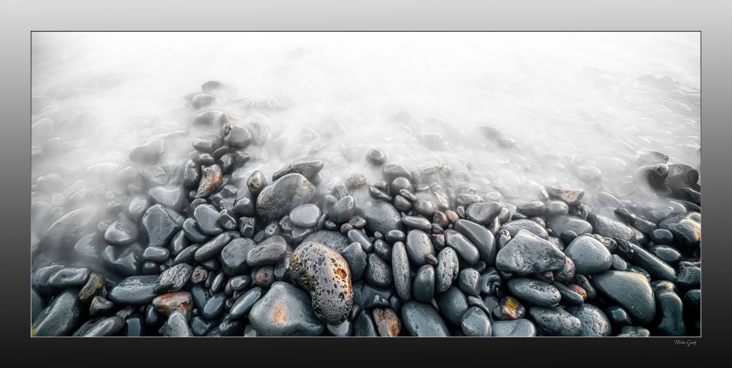Wet gray pebbles with scattered rust-colored markings emerge from pale, softly blurred water.