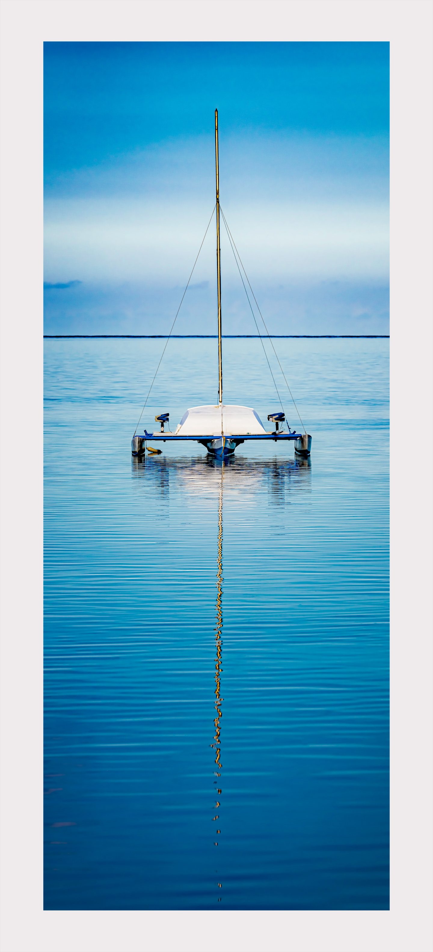 A white and blue trimaran rests on calm water, its tall bare mast casting a long reflection beneath a blue sky.