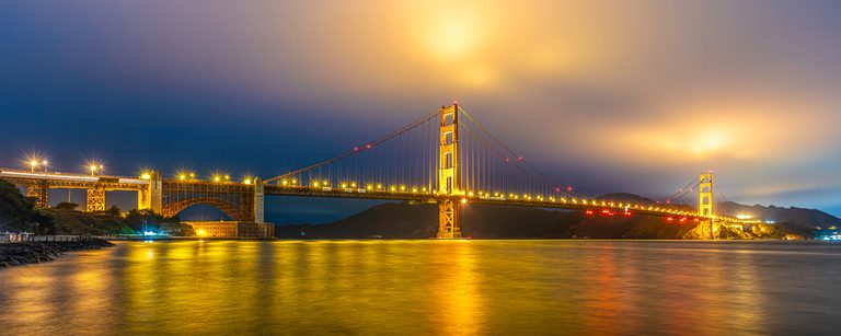 An illuminated suspension bridge crosses dark water, casting golden reflections beneath blue and amber clouds.