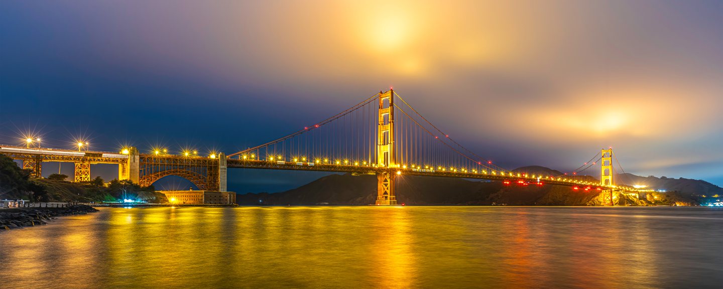 An illuminated suspension bridge crosses dark water, casting golden reflections beneath blue and amber clouds.