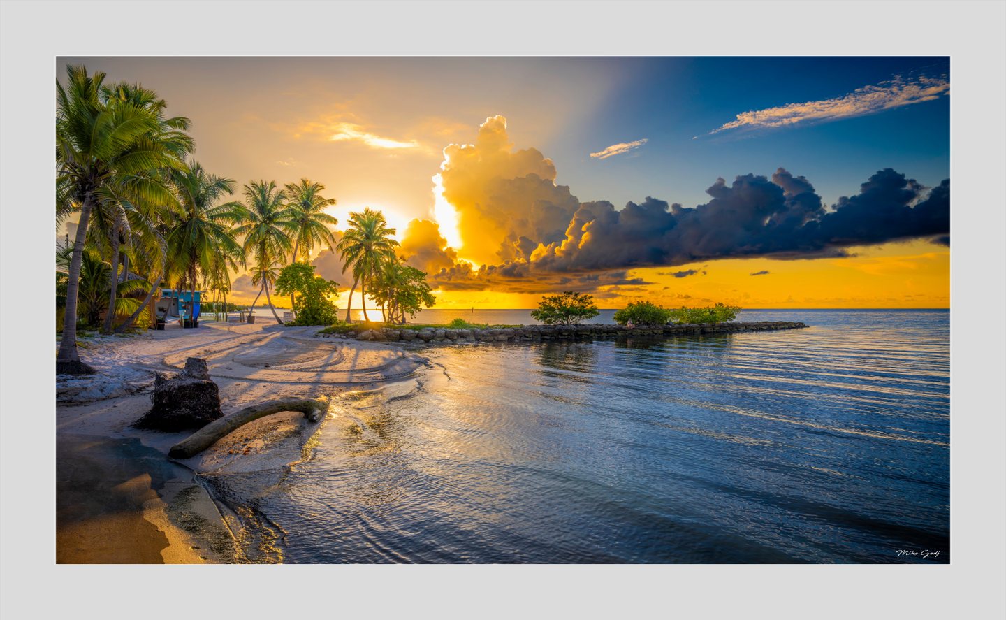 Palm trees line a sandy beach beside calm water beneath towering golden clouds and a deep blue sky.