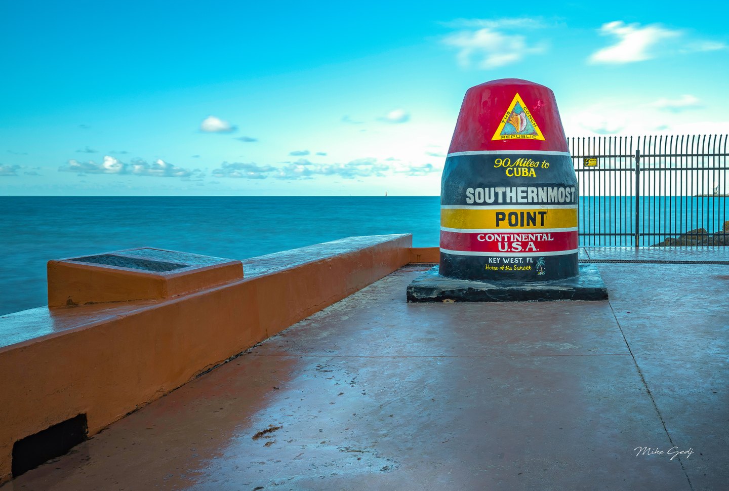 Key West’s red, yellow, and black Southernmost Point marker stands beside a seawall and fence overlooking blue ocean.