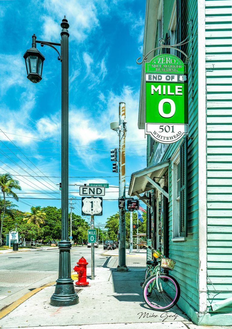 Mile zero signs, a tall streetlamp, and a red hydrant line a sunny sidewalk beside a mint building and bicycles.