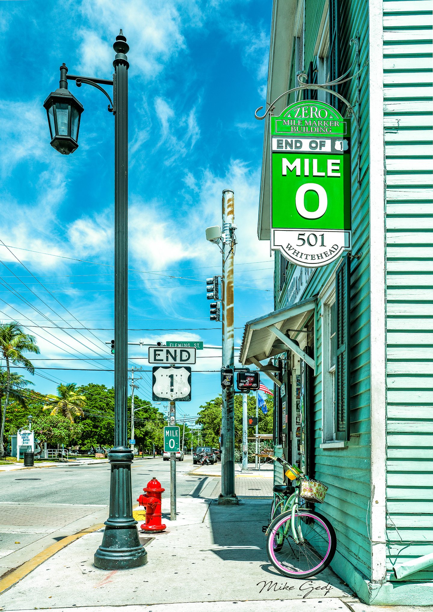 Mile zero signs, a tall streetlamp, and a red hydrant line a sunny sidewalk beside a mint building and bicycles.