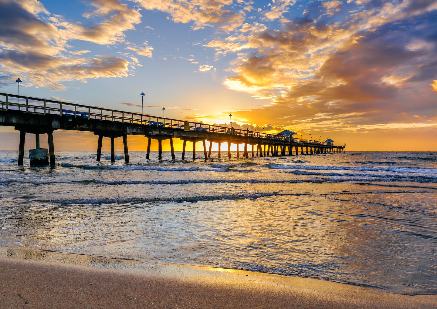 A long pier extends over gentle ocean waves beneath golden clouds, with sunlight reflected on wet sand.
