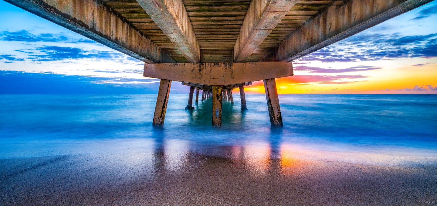 Weathered pier beams and pillars recede over blue ocean water, with orange horizon light reflected on wet sand.