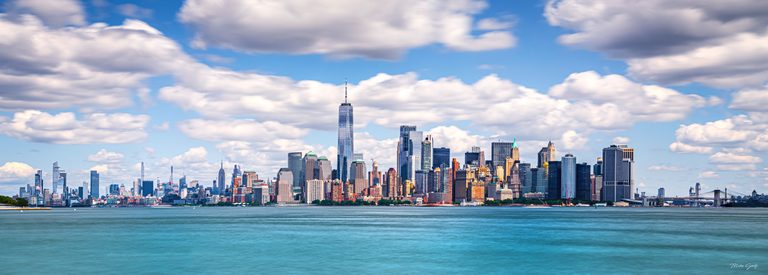 A dense waterfront skyline rises above blue-green water beneath broad white clouds, with a tall spired tower near center.