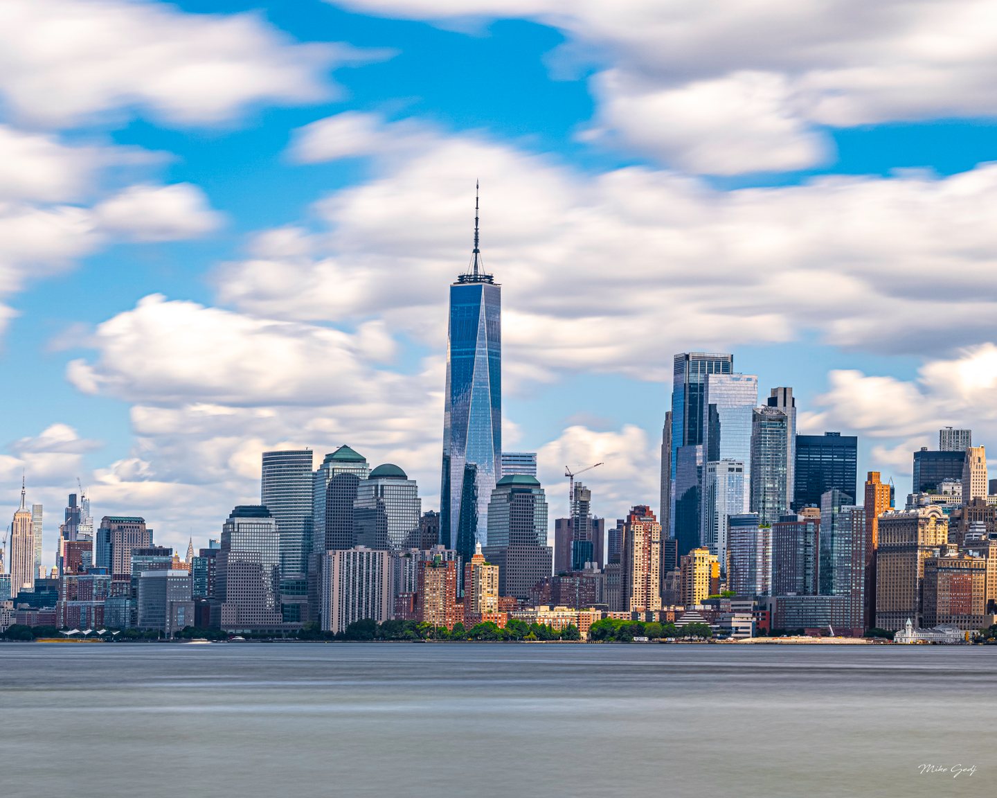 A dense waterfront skyline with a tall glass tower and spire stands beneath white clouds above smooth gray-blue water.