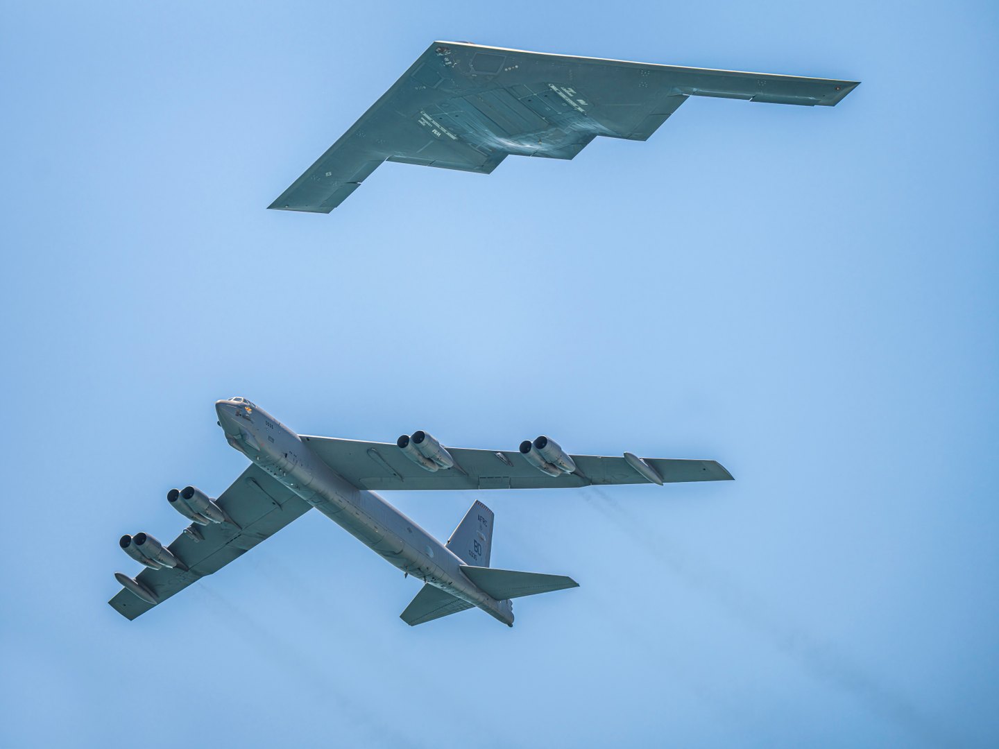 A B-2 stealth bomber and a B-52 bomber seen from below against a clear blue sky.