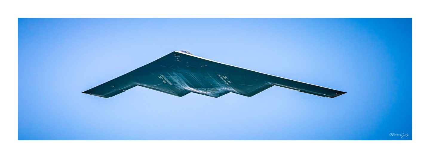 A dark flying-wing stealth bomber seen from below against a clear blue sky, sunlight tracing its angular edges.